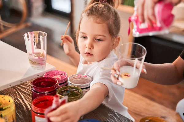 Serious little girl stretching to the paints while drawing pictures on the canvas — Stock Photo, Image