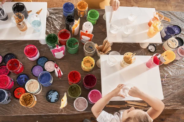 Linda niña preescolar de pie en la mesa y dibujo sobre lienzo con pinturas de diferentes colores —  Fotos de Stock