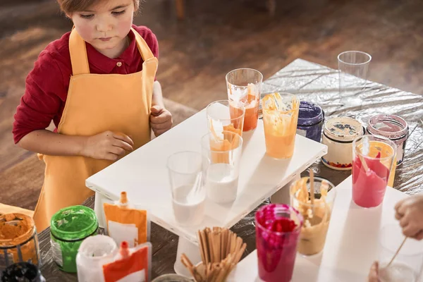 Smart male kid looking attentively while drawing with paints on the canvas and standing at the table — Stock Photo, Image