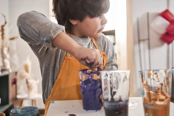 Niño de pie con la cara seria y poniendo sus manos en la taza de plástico con pinturas violetas —  Fotos de Stock