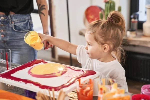 Child girl holding glass with orange paints while standing in front of the table with canvas and drawing — Stock Photo, Image