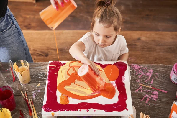 Child blonde girl pouring fresh orange paints into the canvas while creating her own masterpiece — Stock Photo, Image