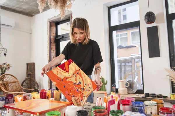 Ženská malířka maluje ve svém pokoji, zatímco stojí u stolu s připravenou abstraktní malbou v rukou — Stock fotografie
