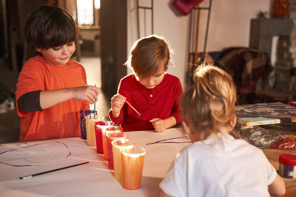 Tres niños felices de pie en la mesa debajo de la lámpara y dibujos con pinturas acrílicas — Foto de Stock