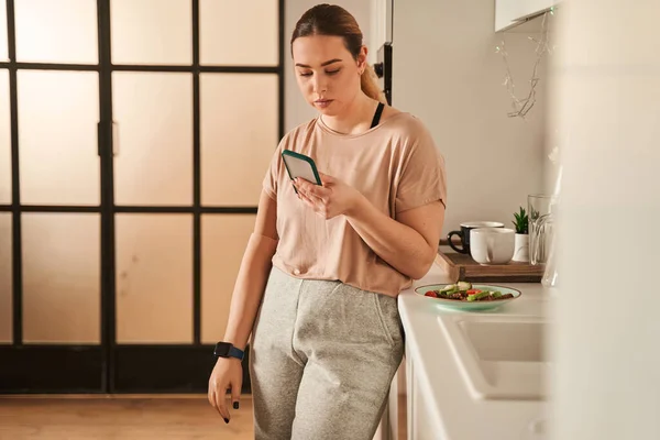 Bella donna serena con mano protesi utilizzando il suo smartphone durante la cottura in cucina — Foto Stock