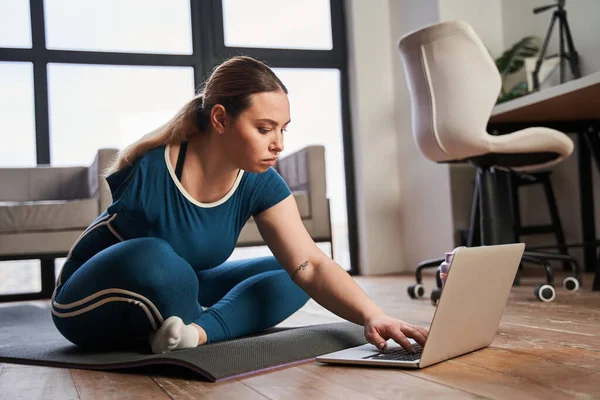 Donna con arto amputato seduto al tappetino yoga e digitare qualcosa al computer portatile — Foto Stock