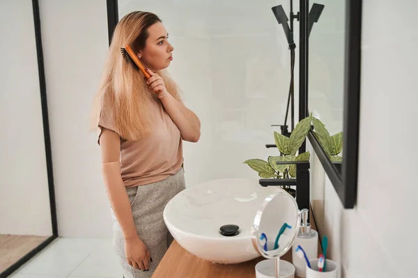 Donna con braccio protesico guardando il suo riflesso nello specchio e spazzolandosi i capelli lunghi — Foto Stock