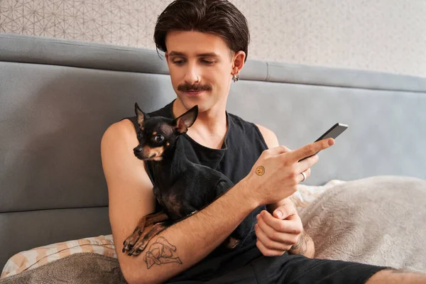 Hombre con bigote sentado en la cama y mirando a su perro mientras usa el teléfono inteligente — Foto de Stock