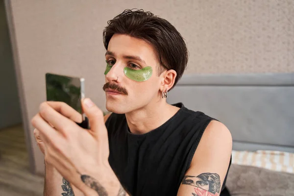 Chico metrosexual con bigote mirando su reflejo mientras se pone máscara debajo de los ojos — Foto de Stock