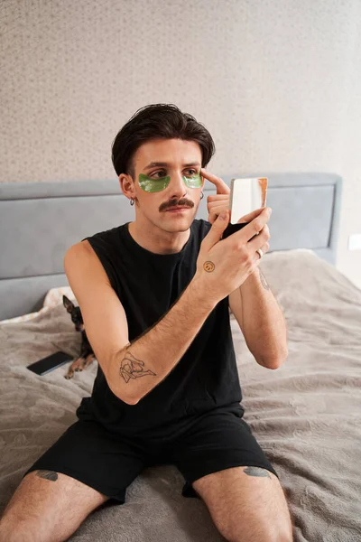 Hombre sentado en la cama y aplicando parches en los ojos mientras sostiene el espejo de la mano — Foto de Stock