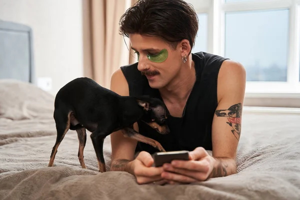 Brune tatouée homme allongé sur le lit avec son smartphone et regardant son chien — Photo