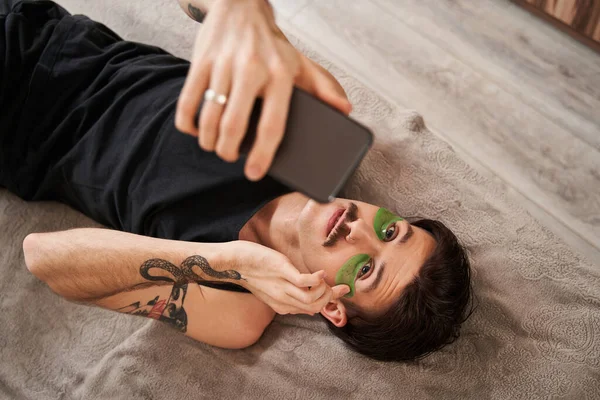 Hombre morena con parches debajo de los ojos acostado en la cama y haciendo selfie en el teléfono inteligente — Foto de Stock