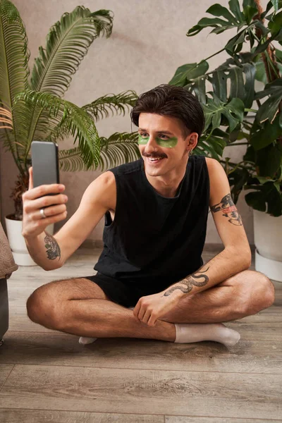 Hombre con parches debajo de los ojos sentado en casa y haciendo selfie en el teléfono inteligente mientras se relaja — Foto de Stock