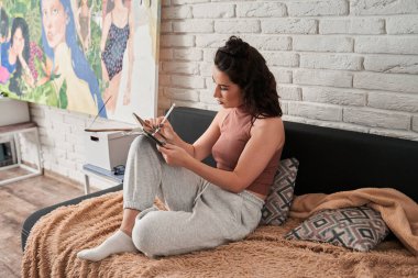 Kanepede otururken konsantre yüzüyle tablete resim çizen esmer kadın.