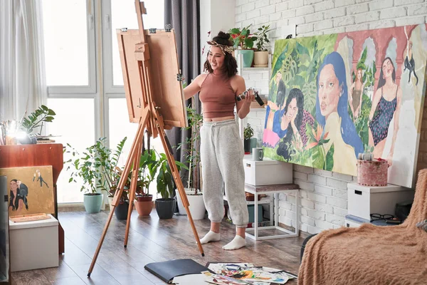 Mulher incrível concentrou-se em pintar enquanto estava de pé e sorrindo na frente do cavalete em seu apartamento — Fotografia de Stock
