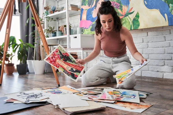 Mujer artista mirando sus obras de arte con cara seria mientras está sentada en el suelo en casa o en el estudio —  Fotos de Stock