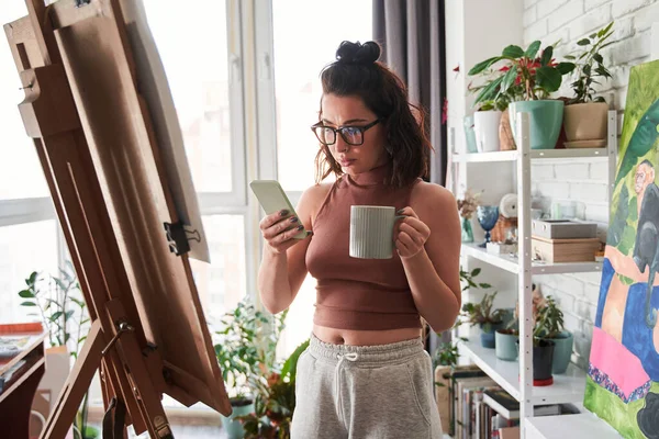 Nő művész kávézás és nézi az okostelefon, miközben időt tölt közelében a festőállvány — Stock Fotó