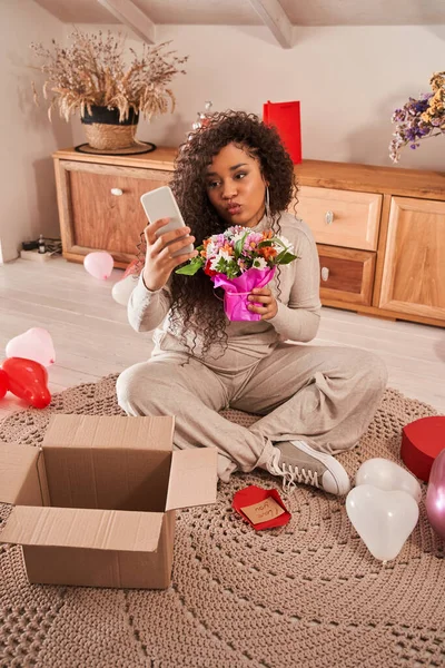 彼女のボーイフレンドからの花でポーズをとりながら、スマートフォンで自分自身を撮影多人種の女性 — ストック写真