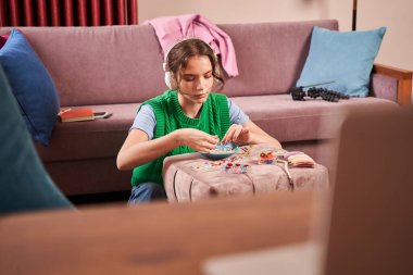 Evde el yapımı mücevher yapan ve büyük kulaklıklarla müzik dinleyen bir genç.