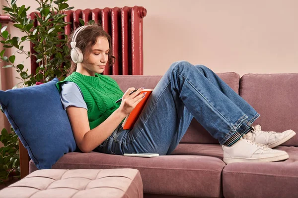Egy tinédzser lány fülhallgatóban ül a kanapén, és ír valamit a füzetébe. — Stock Fotó