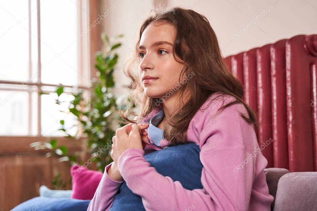 Upset girl frustrated by problem with relationships, sitting on couch and embracing pillow