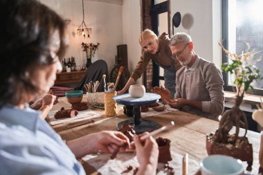 Zanaatkar kadın öğretmen ve arkadaşıyla bankta oturup çömlek atölyesinde kil şekillendiriyor.