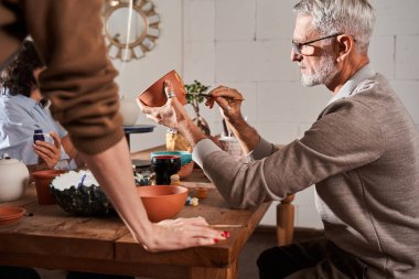 Kil tenceresinde boyayla çizim yapan gözlüklü bir adam ana sınıfta oturuyor.