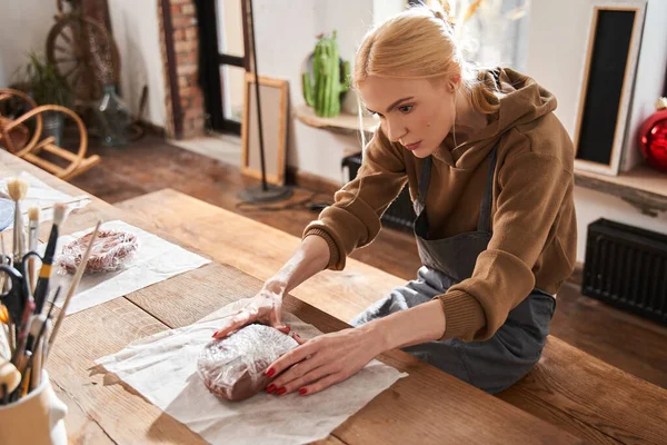 Female artist wearing apron sitting at the bench and looking at the clay with serious face — Stock Photo, Image