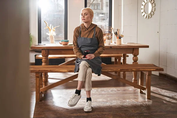 Umělec žena sedí na lavičce s šálkem kávy a dívá se pryč se zasněnou tváří — Stock fotografie