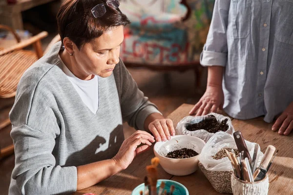 Dojrzała kobieta patrząc na kawałek glazury podczas przygotowywania się do malowania płyty gliny w warsztacie — Zdjęcie stockowe