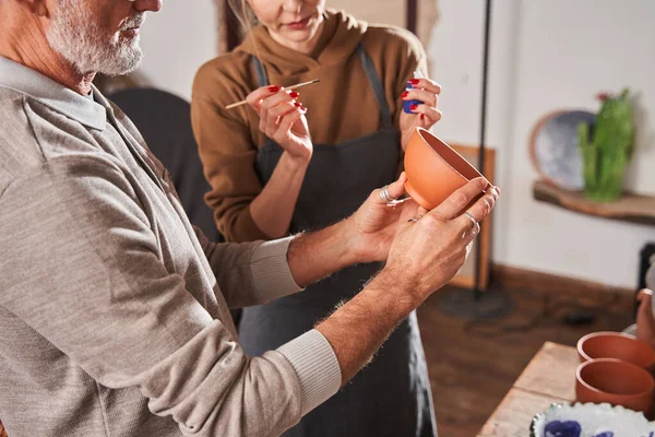 Female artist in apron holding brush and paints for plate and explaining — Stockfoto