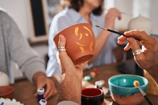 Senior man holding clay pot and brush while drawing patterns on it at the loft studio — Stockfoto