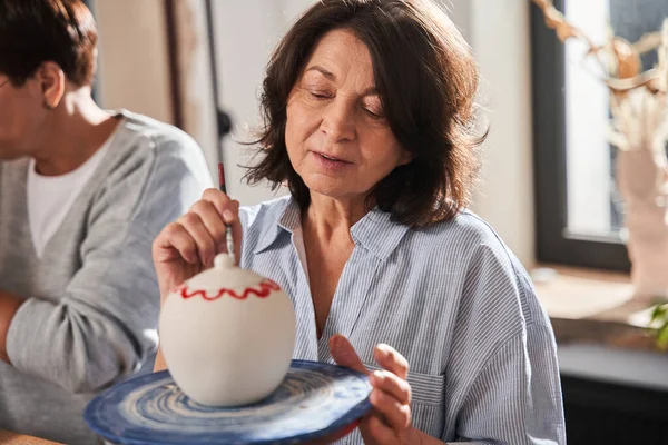 Inspired woman looking at her clay pot and drawing patterns on it — Stock Photo, Image