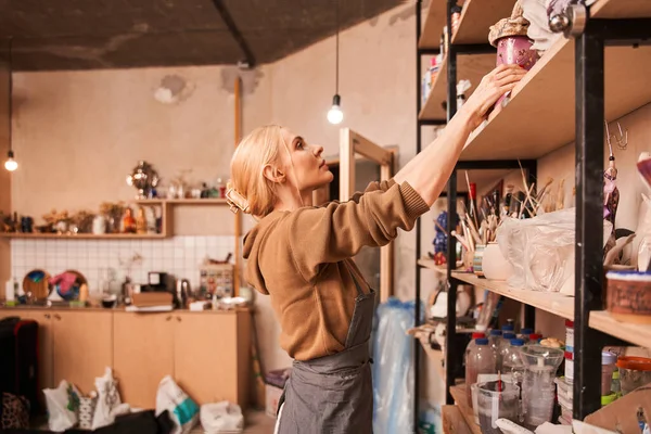 Female artist putting jar at the shelves while preparing drawing at her art space — Stock fotografie