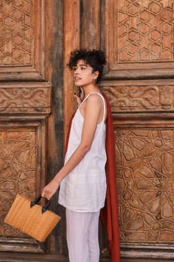 Beautiful brunette woman in fashionable clothes posing near wooden door with bag. Female fashion and summer concept
