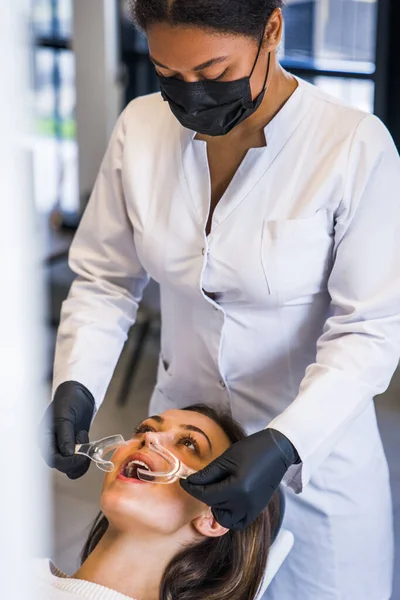 Vertical shot of multiracial woman dentist applies tray for whitening to female patient teeth in modern dental clinic. Teeth whitening in a dental clinic concept