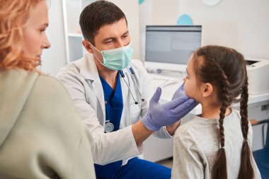 Koruyucu maske takan doktor klinikte liseli kızın boğazını okşuyor, hasta kız çocuk doktorunu ziyaret ediyor. Soğuk algınlığı, koronavirüs, çocuk konseptinde bademcik iltihabı.