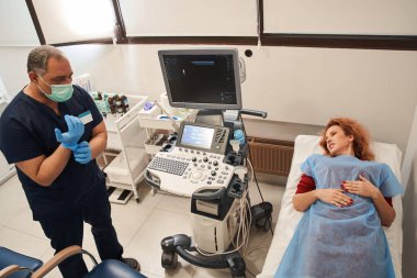 Üst düzey doktorun koruyucu eldivenlerini giyerken kadın hastayla konuşurken ve ultrason muayenesine hazırlanırken yüksek açılı görüntüsü.