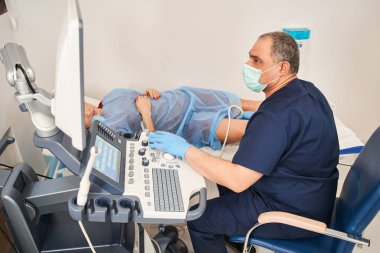 Damarların ultrasonu. Hastanedeki ultrason soruşturmasının yüksek açılı görüntüsü. Yaşlı adam kadın hastanın bacaklarını muayene ediyor ve ekrana bakıyor.