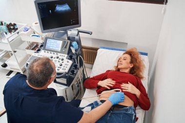 Olgun doktorun özel bir cihaz tutarken ve kadın karnını incelerken yüksek açılı görüntüsü. Kadın modern teknoloji kliniğinde ultrason sensörü üzerinde tıbbi muayeneden geçiyor.