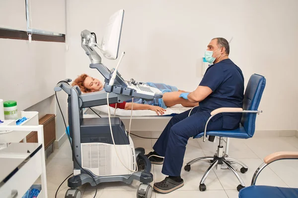 Üniformalı bir doktor, teşhis cihazıyla kadın bir hastanın bacaklarındaki cihazı kullanarak ultrason yapar. Cerrah, filebolog. Moder Kliniği 'nde çalışıyor. Hastanede tıbbi aletler