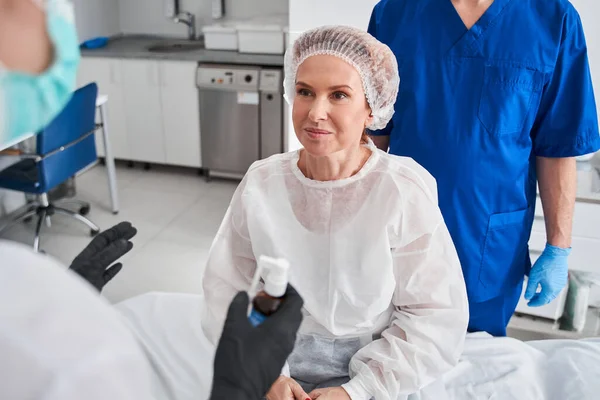 Happy woman sitting at the hospital bed and preparing to the gastroscopy. Doctor and nurse at the background. Stock photo