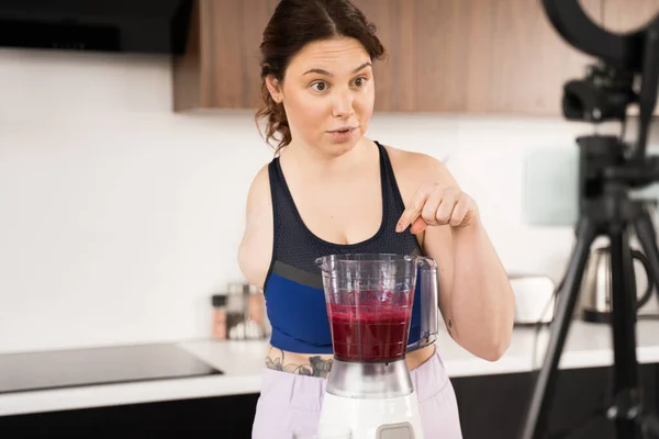 Waist up portrait view of the woman without hand standing in front of the blender with smoothie and telling about it while recording video blog about healthy lifestyle