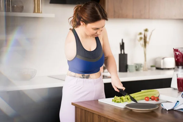 Woman with amputee arm chopping celery at the board and preparing to the healthy dinner while cooking at the kitchen. Food and vegetarian lifestyle concept