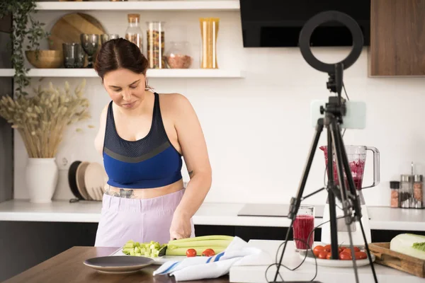Woman with amputee arm chopping celery at the board and recording video blog about healthy lifestyle while cooking salad at the kitchen. Food and vegetarian lifestyle concept