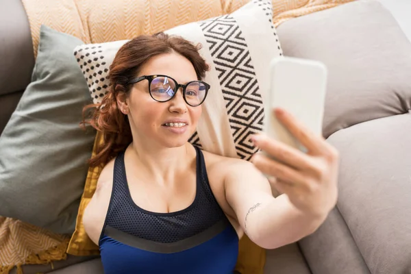 High angle view of the woman with amputee arm wearing glasses laying at the couch and taking selfie at her mobile phone. Technologies and people concept
