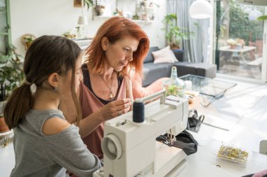 Concentrated hardworking skilled female designer sewing new clothes while sitting at her workplace at home with daughter