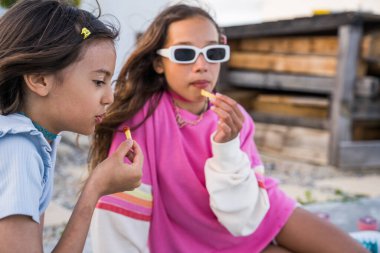 Charming girls chews a fried potato with appetite and chatting with each other at the street. Cheerful girl eating raspberries from her fingers