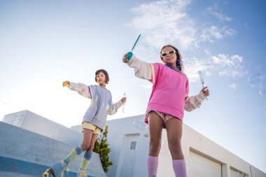 Low angle view of the little caucasian girlfriends in vivid clothes with soap bubbles at hands riding at the roller skates. Friendship concept. Stock photo