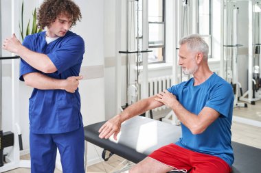 Young physical therapist showing to senior patient how to do while working with him at the rehabilitation centre. Stock photo